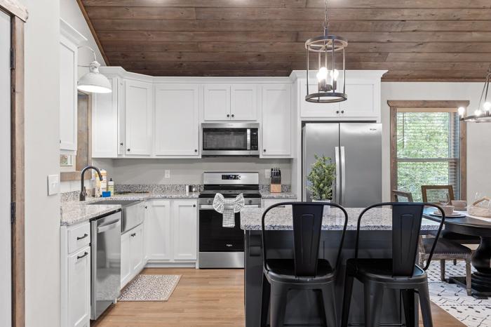 A modern kitchen with white cabinets, stainless steel appliances, a wooden ceiling, and a dining area with black chairs.