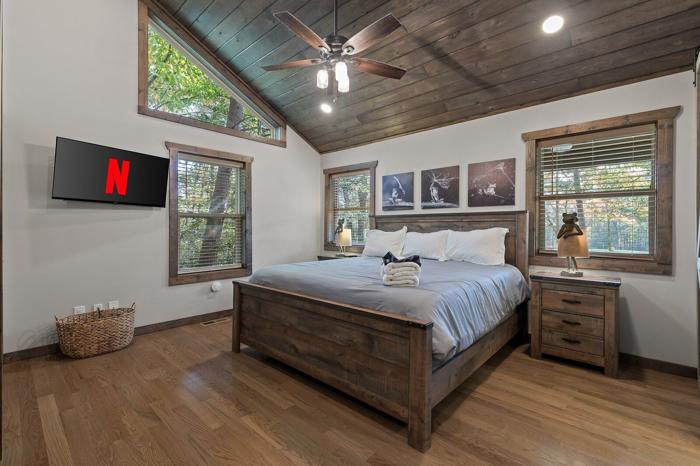 A bedroom with a wooden ceiling fan, a bed with a grey bedspread, a television mounted on the wall, and a window with blinds.