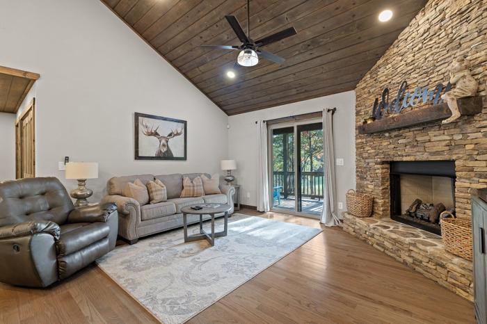 A living room with a stone fireplace, a ceiling fan, a sofa, a leather chair, and a large window.