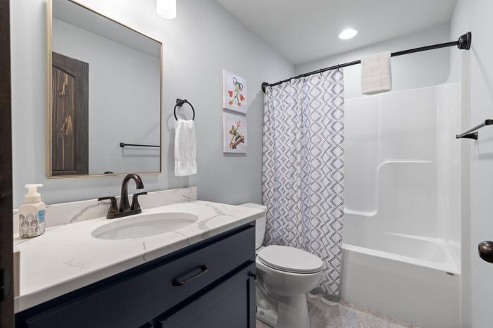 A bathroom with a white sink, dark cabinets, a toilet, a shower with a patterned curtain, and a mirror with a wooden frame.