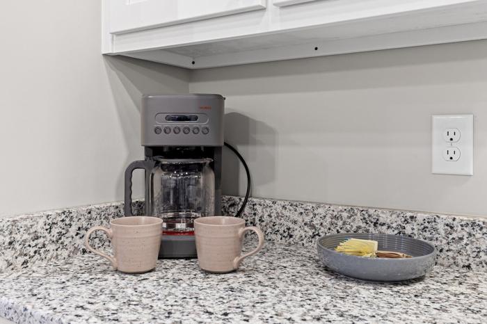 A coffee maker on a granite countertop with two mugs and a bowl of sugar and creamers.