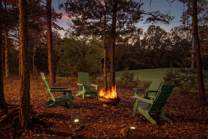 A campfire with two green Adirondack chairs around it, set in a wooded area with trees and a grassy field in the background.
